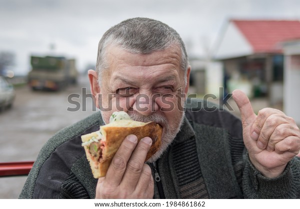 Portrait of hungry Caucasian senior driver\
eating lyulya kebab in lavash near his\
car