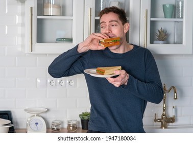 Portrait Of Hungry Guy, Young Man In Pyjama Is Eating Tasty Sandwich, Biting Burgers At Home At Kitchen With Gusto At Breakfast In A Hurry. Unhealthy Fast Food.