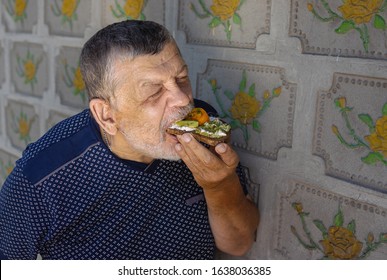 Portrait Of Hungry Senior Eating Vegan Sandwich With Cucumber, Yellow Tomato And Guacamole While Standing Against Tiled Wall