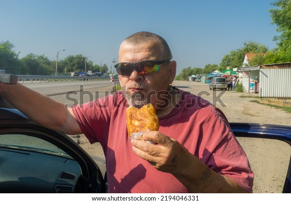 Portrait of hungry Ukrainian senior driver taking\
patty near his car while standing near Vasylivka, Zaporizhzhia\
Oblast, Ukraine