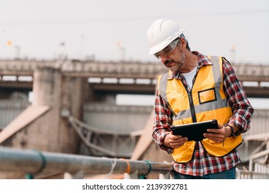 Portrait Of Hydropower Engineer Wearing Safety Jacket And Hardhat With Tablet Working At Outdoor Field Site That Have Water Spillway  Of Hydro Power Dam Electrical Generator At The Background.