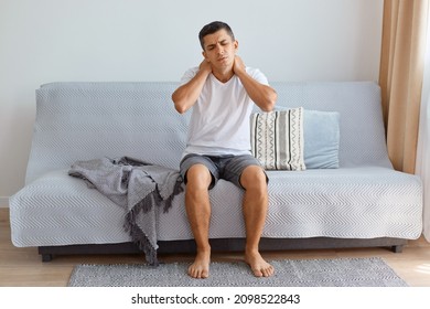 Portrait of ill unhealthy man wearing white T-shirt sitting on cough in living room, touching or massaging his painful neck, uncomfortable sleeping conditions, vertebral column injury. - Shutterstock ID 2098522843