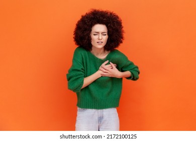 Portrait Of Ill Woman With Afro Hairstyle Wearing Green Casual Style Sweater Having Hear Attack, Frowning From Strong Pain, Needs Hospitalization. Indoor Studio Shot Isolated On Orange Background.