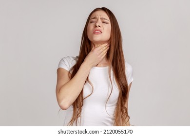 Portrait of ill woman with brown hair standing touching neck and frowning from pain, suffering sore throat, flu symptom, wearing white T-shirt. Indoor studio shot isolated on gray background. - Powered by Shutterstock