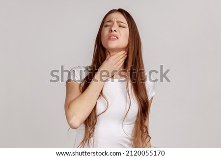 Portrait of ill woman with brown hair standing touching neck and frowning from pain, suffering sore throat, flu symptom, wearing white T-shirt. Indoor studio shot isolated on gray background.