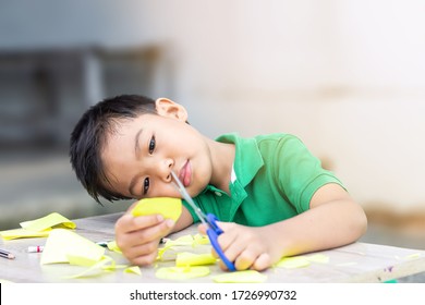 Portrait Image Of 5-6 Years Old Asian School Kid Learning To Cutting Color Paper By Scissors,Bored Child Boy Stay At Home During Quarantine And Social Distancing,Home Schooling, Distance Education.