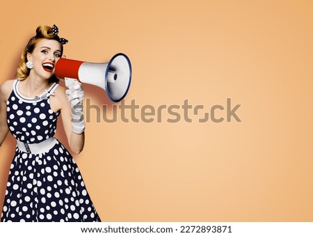 Portrait image of beautiful woman holding mega phone, shout, saying, advertising. Pretty girl in black pin up style dress with megaphone loudspeaker. Isolated latte beige background. Big sales ad.