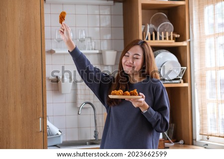 Portrait image of a beautiful young asian woman holding fried chicken and dancing in the kitchen at home