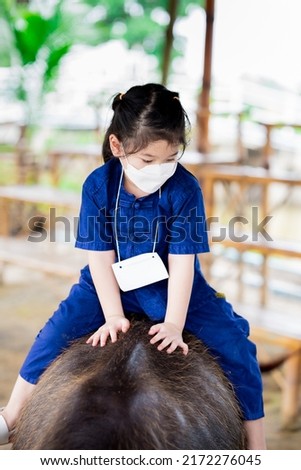 Portrait image child 5 year old. Cute Asian girl is riding on back of black buffalo. Children get close to farm animals. Learning rural lifestyle. Lessons outside classroom. Kid wear blue farmer dress
