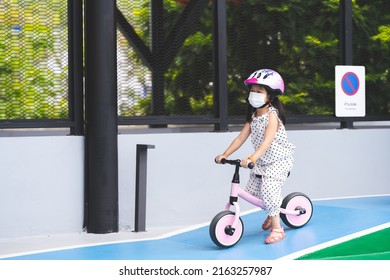 Portrait Image Child 5 Years Old. Cute Asian Girl Wearing White Face Mask Is Riding Pink Plow Bike Down Small Hill In Training Ground, Traffic Sign With Thai And English Writing No Parking And Symbol.