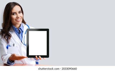 Portrait Image Of Happy Smiling Female Doctor Showing Tablet Pc Ipad Touchpad With Blank Copy Space Area For Slogan Or Text, Over Grey Colour Background. Medical Center Centre Ad Concept.