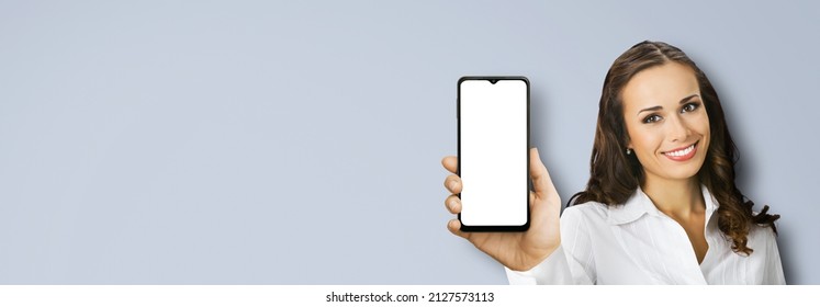 Portrait Image Of Happy Smiling Young Woman In Confident White Cloth Holding Showing Smartphone Cell Phone Mobile With Empty Mock Up Screen, Isolated Over Grey Background. Consulting Assistance