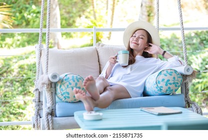 Portrait image of a young woman with hat drinking coffee while relaxing and laying down on swing sofa 