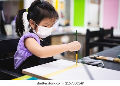 Portrait Imaged Child Aged 5 Years Old. Kindergarten Kid Learning Art Water Color. Children Wearing White Medical Face Mask To Reduce Spread Of Virus. Girl In Classroom Doing Craft With Paintbrush.