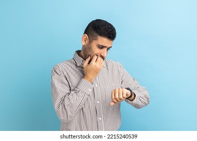 Portrait Of Impatient Businessman Pointing Finger On His Wrist Watch With Nervous Face, Time Is Out, Deadline, Wearing Striped Shirt. Indoor Studio Shot Isolated On Blue Background.