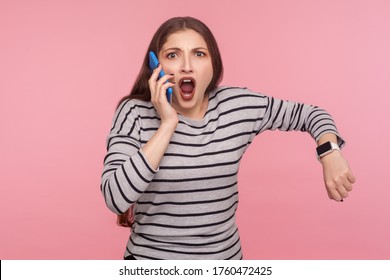 Portrait Of Impatient Woman In Striped Sweatshirt Talking On Mobile Phone And Looking With Shocked Angry Expression, Showing Wrist Watch, Mad About Late Time. Studio Shot Isolated On Pink Background