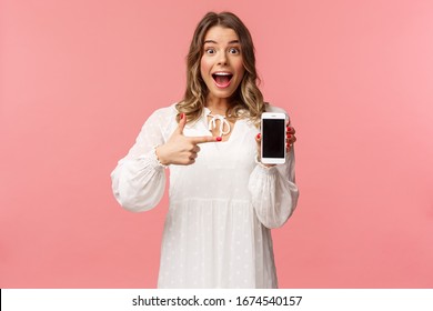 Portrait Of Impressed, Excited Young Blond Woman Showing Something Awesome On Display, Pointing Mobile Phone Screen And Smiling Astonished, Brag With Her Recent Match On Dating App