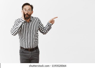 Portrait Of Impressed And Thrilled Gay Man With Beard And Moustache, Folding Lips In Curious Emotion And Pointing Right, Holding Palm On Cheek, Being Surprised And Shocked Person Showed Up On Party