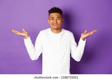 Portrait Of Indecisive African-american Guy In White Sweatshirt, Raise Hands Sideways And Shrugging Clueless, Standing Confused Against Purple Background