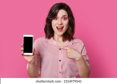 Portrait of independent, excited girl in pink, striped shirt with round buttons holding white mobile phone with black screen in fingers, and her second hand points to switched off smartphone. - Shutterstock ID 1314979541