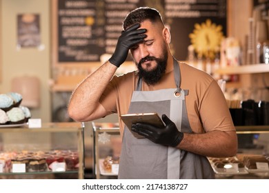 Portrait of Indian barista in grey apron using digital tablet standing at the modern cafe. Frustrated cafe owner using digital tablet has business problems. Upset bearded man receiving online orders - Powered by Shutterstock