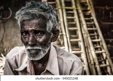 Portrait of Indian elder man with traditional bindi as a third eye, white beard and bamboo ladders on the background in Mysore, Karnataka, India. Rural village dark skin old man, concerned expression