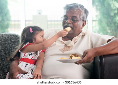 Portrait Of Indian Family At Home. Grandchild Feeding Butter Cake To Grandparent. Grandfather And Granddaughter. Asian People Living Lifestyle.