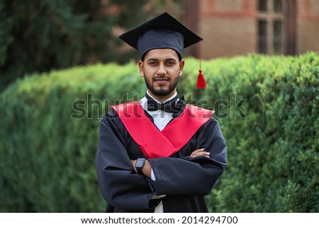 Portrait of indian handsome male graduate in graduation robe with crossed arms and blured background.