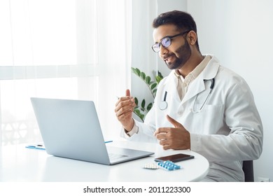 Portrait of indian man doctor talking to online patient on laptop screen sitting at clinic office desk giving online consultation for domestic health treatment. Telemedicine remote medical appointment