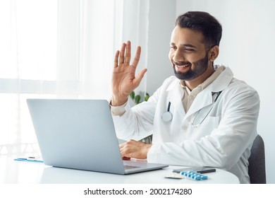 Portrait Of Indian Man Doctor In Uniform Waving Hand Greeting Online Patient On Laptop Screen Sitting At Hospital Office Desk. Online Consultation Service, Telemedicine Application For Sick People