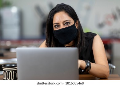 Portrait Of An Indian Woman Wearing Covid 19 Protection Mask And Working On Her Laptop, Sitting In An Office Cafeteria, Coffee Shop, Casual Work Environment.