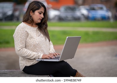 Portrait Of Indian Woman Working On Computer At Park With Cars In Background. South Asian Indian Woman Typing On Laptop Outdoor.