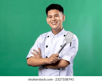 A portrait of an Indonesian Asian man wearing a chef's uniform, seen crossing his arms and holding a spatula with one hand, isolated with a green background. - Powered by Shutterstock