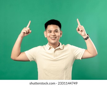 A portrait of an Indonesian Asian man wearing a yellow shirt, looking happy with his hand pointing upwards, isolated on a green background. - Powered by Shutterstock