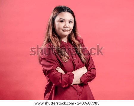 Portrait of an Indonesian Asian woman, wearing a red dress, confidently posing with her hands folded, isolated against a pink background.