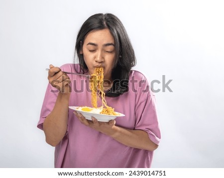 Portrait of an Indonesian Asian woman, wearing a pink shirt, happily eating instant noodles, isolated against a white background.