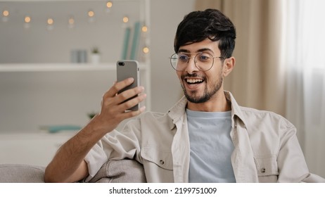 Portrait Indoor Male Arabic Guy Unshaven Man With Glasses Sitting On Couch At Home Chatting Online Using Mobile Phone Reads News Scrolling Browsing Net Typing In Smartphone Surprised Answers Message