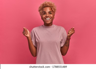 Portrait Indoors Of A Dark-skinned Girl With A Short Haircut Who Finally Got What She Really Wanted. Woman In Casual Clothes Stands On A Background Of A Pink Wall And Is Sincerely Happy.