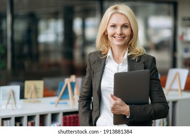 Portrait of influential elegant businesswoman with blonde hair in stylish formal suit. Happy successful middle aged lady looks directly at the camera, friendly smiling, holding laptop in arms