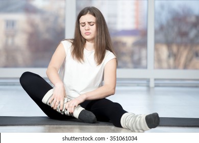 Portrait of injured unhappy fit young beautiful woman sitting in sports club, touching her ankle after working out in class, suffering with closed eyes, feeling pain, full length