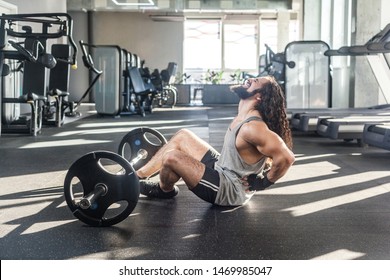 Portrait Of Injury Young Adult Man Athlete With Long Curly Hair Working Out In Gym, Sitting On Floor And Have Strong Hurt Problem With Back, Spasm Painful. Gripping Spinal With Hand, Screaming. Indoor