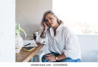 Portrait of intelligent Caucasian female student looking at camera during time for learning and studying in cafe interior, clever hipster girl with education textbook and mobile phone posing indoors