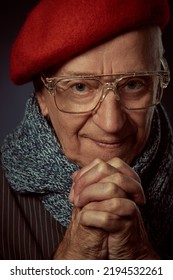 Portrait Of An Intelligent Elderly Man In A Beret, Scarf And Glasses Standing With Folded Hands In Front Of Him And Smiling. Old Age Concept. Dark Background.