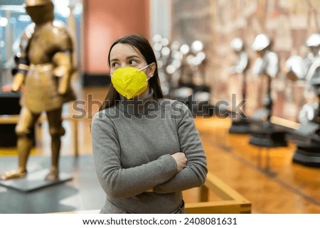 Portrait of interested adult brunette wearing protective face mask visiting exhibition of medieval armor in armory of historical museum during coronavirus pandemic