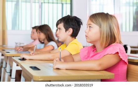 Portrait Of Interested Blonde Tween Girl Studying In Classroom, Listening To Schoolteacher And Writing In Notebook