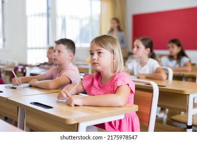 Portrait Of Interested Blonde Tween Girl Studying In Classroom, Listening To Schoolteacher And Writing In Notebook