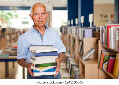 Portrait of interested older man standing in library with pile of books in hands