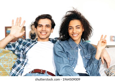 Portrait Of Interracial Friendly Happy Young Family Couple Sitting On Sofa, Stylishly Dressed, Looking Waving Hands At The Camera, Smiling, Spend Time Together. Relationship Between Multiethnic People