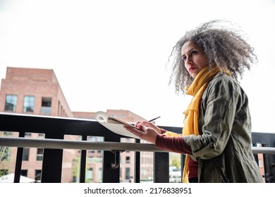 Portrait Of Iranian Mature Artist Woman Making A Draw On A Sketchbook In London Outside. She Is Leaning On A Banister And Wearing A Yellow Scarf And Army Jacket. Curly Grey Hair.