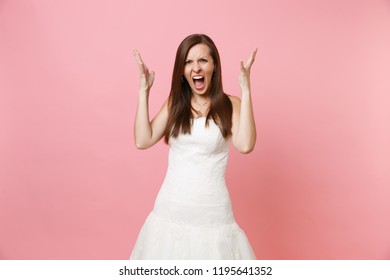 Portrait of irritated angry bride woman in beautiful white wedding dress stand screaming spreading hands isolated on pink pastel background. Wedding celebration concept. Copy space for advertisement